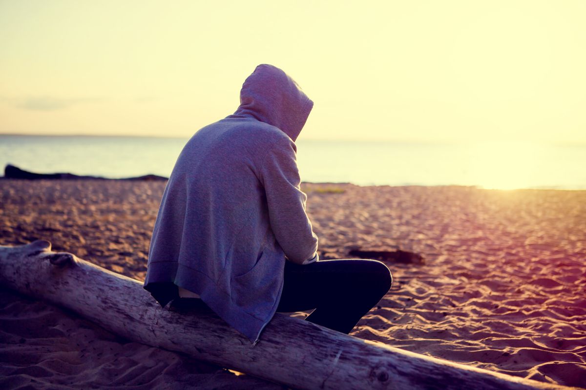 Person in hoodie sitting on a log at the beach and looking at the water