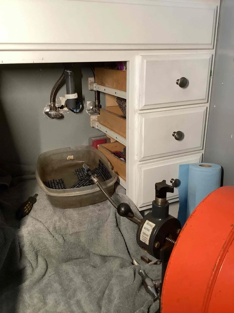 Plumbing tools and a snake machine under a sink with open drawers, surrounded by towels and a roll of blue paper towels.
