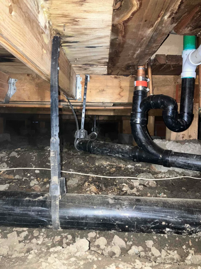 Under-floor view showing new plumbing pipes installed for repiping, secured to wooden beams, amid dirt and debris.