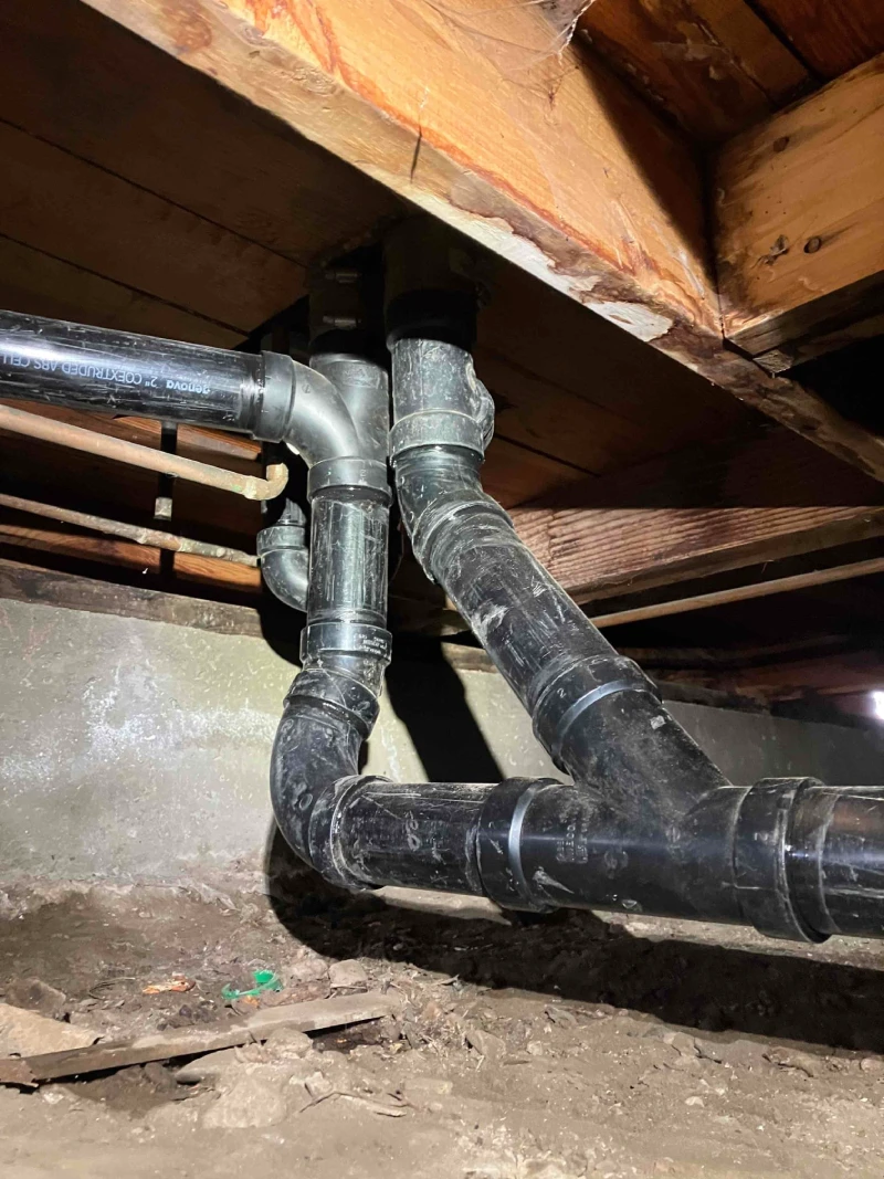 New plumbing pipes installed under a wooden floor, showcasing recent repiping work in a dimly lit basement area.