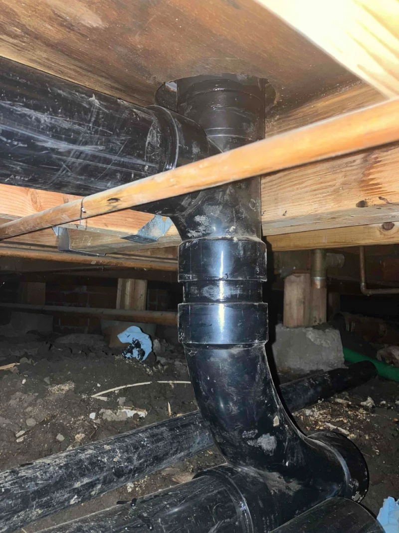 Black pipes under a wooden floor show recent plumbing work, highlighting the repiping process in a crawl space setting.