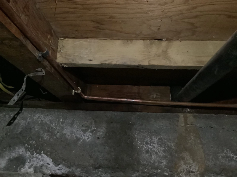 Copper pipes under a wooden floor show a recent water line repair in a dimly lit basement, highlighting the plumbing work.