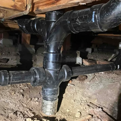 New black pipes installed under a wooden floor highlight recent plumbing repiping work in a dimly lit crawl space.