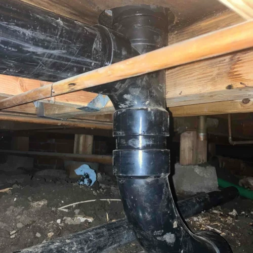 Black pipes under a wooden floor show recent plumbing work, highlighting the repiping process in a crawl space setting.