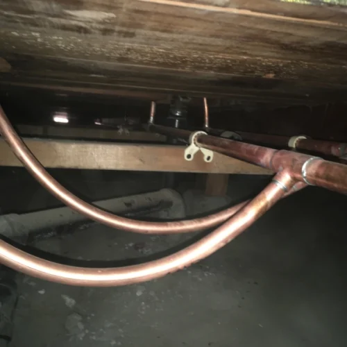 Copper pipes under a wooden floor show recent plumbing repiping work, highlighting new installations in a dimly lit space.
