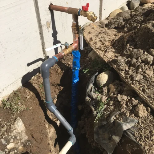 Plumbing repiping project shows exposed pipes and fittings next to a building, with soil and rocks surrounding the trench.
