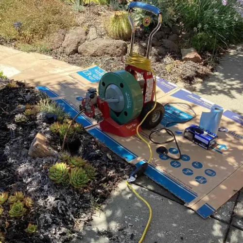 Sewer repair equipment set up in a garden for sewer services, surrounded by plants and tools on a sunny day.