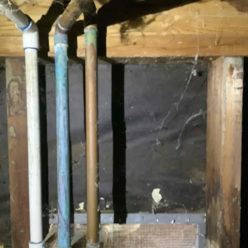 Pipes under a wooden floor show signs of corrosion, indicating a need for water line repair in the dimly lit space.