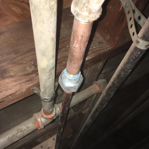 Old pipes under a wooden floor show signs of water line repair, with new fittings and joints visible against the aged metal.