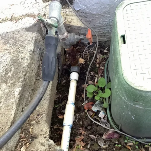 "Outdoor water line repair scene showing exposed pipes and valves near a wall, surrounded by soil and leaves.